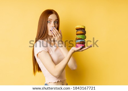 Shot of slim nice surprised caucasian redhead woman in casual t shirt holding pile of colorful donuts, looking with puzzled expression over yellow background. Bakery, unhealthy food, diet concept.