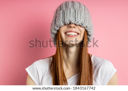 Close up portrait of cheerful happy girl with red long hair having fun, covering eyes with big loop knitted hat, smiling broadly with perfect white teeth isolated over pink background. Royalty-Free Stock Photo #1843167742