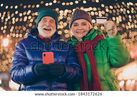 Photo of two people retired friends hold telephone debit card advice online season shopping service wear coat red scarf headwear x-mas evening street park illumination fair outdoors outside