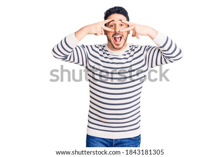 Young hispanic man wearing casual clothes doing peace symbol with fingers over face, smiling cheerful showing victory 