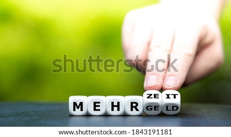 Hand turns dice and changes the German expression "mehr Geld" (more money) to "mehr Zeit" (more time). 