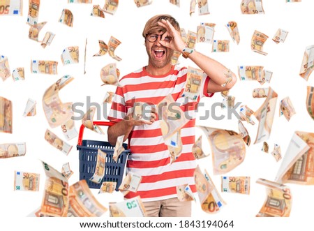 Handsome caucasian man with beard holding supermarket shopping basket smiling happy doing ok sign with hand on eye looking through fingers