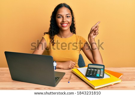 Young african american girl working at the office with laptop and calculator with a big smile on face, pointing with hand finger to the side looking at the camera. 