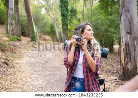 Pensive lady holding camera, looking around and standing on road. Female tourist exploring nature and taking photo of forest. Greenery on background. Tourism, adventure and summer vacation concept