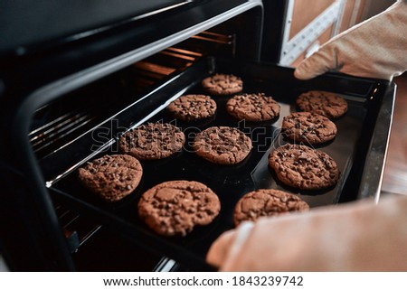 Close up of hands taking out a baking sheet from the oven with cookies with chocolate. Baking process concept Royalty-Free Stock Photo #1843239742