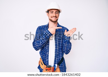 Young handsome man weaing handyman uniform amazed and smiling to the camera while presenting with hand and pointing with finger. 