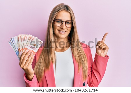 Young blonde woman wearing business style holding mexican pesos smiling happy pointing with hand and finger to the side 