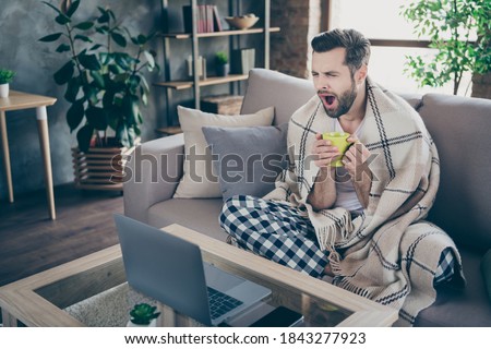 Photo of handsome homey guy sit sofa just wakeup work morning drink coffee yawning energy browsing notebook freelancer remote work stay home quarantine pajama living room indoors