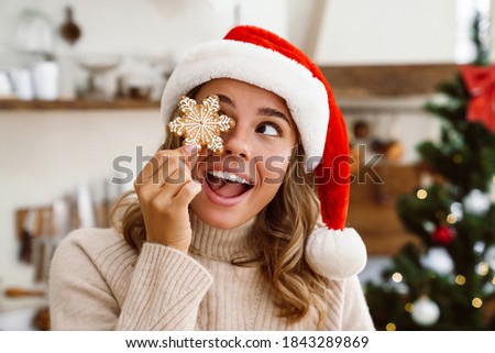 Happy cute woman in santa claus hat making fun with Christmas cookie in cozy kitchen
