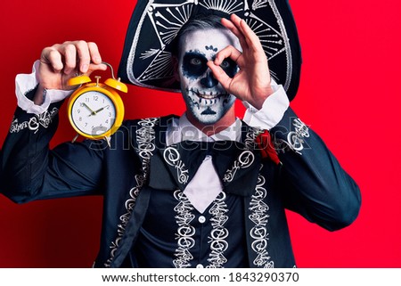 Young man wearing mexican day of the dead costume holding alarm clock smiling happy doing ok sign with hand on eye looking through fingers 