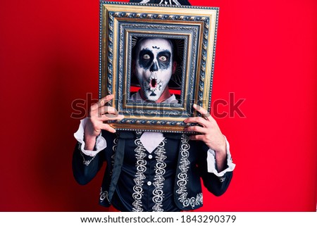 Young man wearing mexican day of the dead costume holding empty frame scared and amazed with open mouth for surprise, disbelief face 