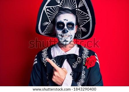 Young man wearing mexican day of the dead costume over red cheerful with a smile of face pointing with hand and finger up to the side with happy and natural expression on face 