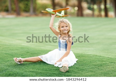 Launch into the air. Happy little girl in dress sitting on the grass field with green toy plane in her hands.
