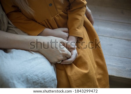 Mom's hug. Daughter and mom are sitting on a chair and mom holds her daughter's hands tightly in her arms. Childhood and motherhood. A daughter in an orange linen dress on her mother's lap. 