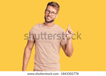 Young caucasian man wearing casual clothes and glasses with a big smile on face, pointing with hand finger to the side looking at the camera. 