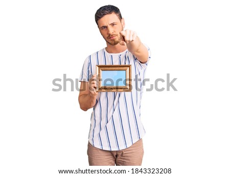 Handsome young man with bear holding empty frame pointing with finger to the camera and to you, confident gesture looking serious 