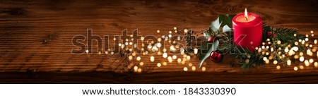 Background for christmas with burning red candle and natural decoration on rustic wooden board. Long horizontal Background with festive golden bokeh lights and space for your text.
