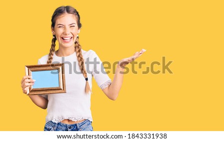 Beautiful caucasian woman with blonde hair holding empty frame celebrating victory with happy smile and winner expression with raised hands 