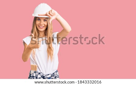 Beautiful caucasian woman with blonde hair wearing hardhat and painter clothes smiling making frame with hands and fingers with happy face. creativity and photography concept. 