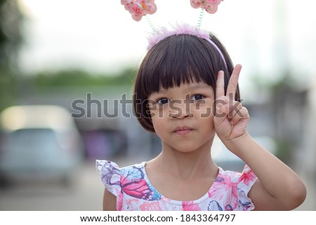 Portrait close up, a 4 to 6 year old Thai Asian kid girl with short hair, wearing a headband on her head, in a sweet dress, pretty face, raising her left hand, raising two fingers beside her head.