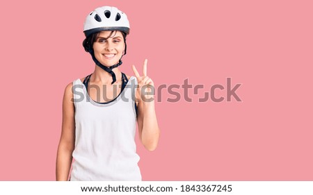 Beautiful young woman with short hair wearing bike helmet smiling with happy face winking at the camera doing victory sign. number two. 