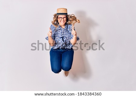 Young beautiful blonde woman wearing glasses and summer hat smiling happy. Jumping with smile on face doing ok sign with thumbs up over isolated white background.