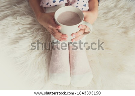 Woman hands holding cup of hot cocoa at home. Top view on girl hands and legs in warm kntitted long socks. Winter and stay at home concept with copy space.