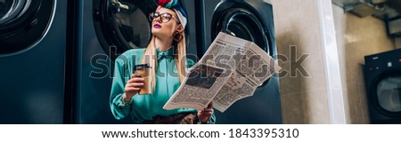 trendy woman in glasses and turban holding paper cup and newspaper in laundromat, banner