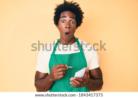 Handsome african american man with afro hair wearing waiter apron taking order afraid and shocked with surprise and amazed expression, fear and excited face. 