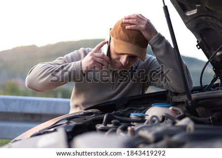 Angry young man waiting a help while sitting near the broken car at the side of road. Roadside assistance concept