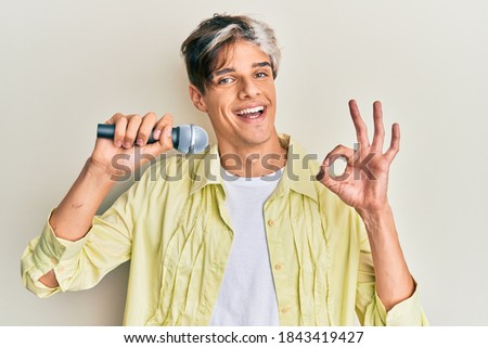 Young hispanic man singing song using microphone doing ok sign with fingers, smiling friendly gesturing excellent symbol 