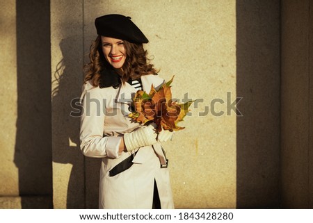 Hello november. smiling elegant woman in beige trench coat with autumn yellow leaves outside in the city in autumn.