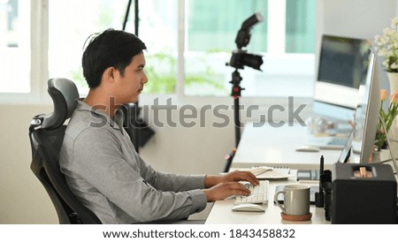 Sideview of photographer editing photos after a photo shoot on desktop with camera and lens placed on table.
