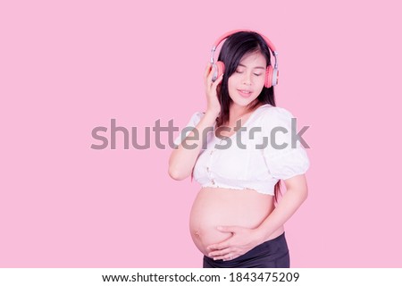 Asian beautiful pregnant woman stands relaxed and enjoys listening to music on headphones connected to the internet