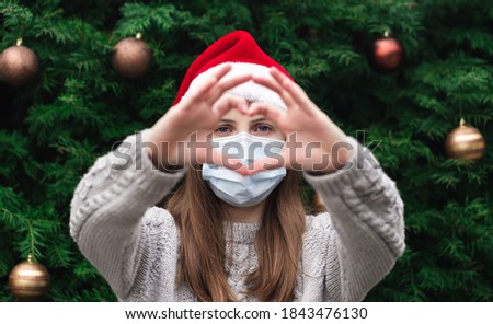 Hand shaped heart love. Close up Portrait of woman wearing a santa claus hat and medical mask with emotion. Against the background of a Christmas tree. Coronavirus pandemic Royalty-Free Stock Photo #1843476130