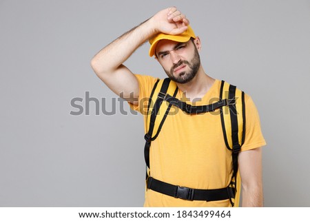 Delivery employee man guy male 20s in yellow cap t-shirt uniform thermal food bag backpack work courier service during quarantine coronavirus covid-19 virus, posing isolated on gray background studio