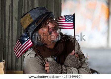 Homeless elderly man wearing hat sitting, smiling and holding flag USA