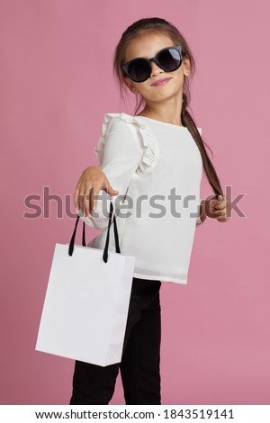 Sale. adorable fashionable little child girl in sunglasses is holding shopping bag on pink background. daughter wants to be like mom