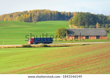 Modern semi-trailer truck riding through green plowed agricultural field with tractor tracks. Forest in the background. Germany. Highway, freight transportation, articulated lorry, logistics, industry