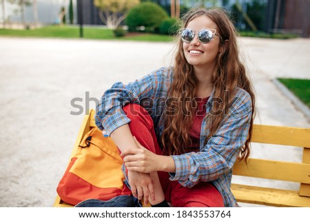 Happy young blonde hipster woman student with backpack looking sitting on bench at university.