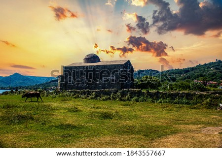 Cow grazing in front of the Greek Church and church in a green field under a cloudy sky.