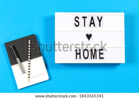 White lightbox with text STAY HOME and notebook on blue background. Coronavirus, Covid-19, online learning, working from home, quarantine, social distancing concept