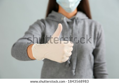 Woman in protective face mask and medical gloves showing thumb up gesture on grey background, closeup