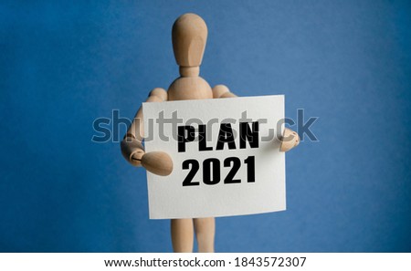 The wooden man holds a white sign with the text PLAN 2021 in his hands. The content of the lettering has implications for business concept and marketing.
