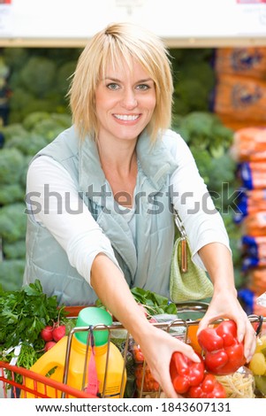 Vertical waist up portrait of a young woman loading red bell pepper into her shopping cart at a grocery store smiles at the camera.