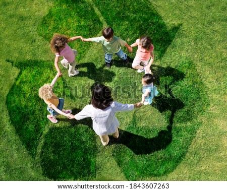 Overhead shot of teacher with five young students in a circle forming a human chain play on a green lawn outdoors with overlay of recycle symbol.