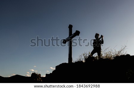 Silhouette of man using camera and standing on the top of a hill or mountain next to Christian cross.Traveler taking photos of landscape below.Twilight blue sky in the background,hiking travel concept