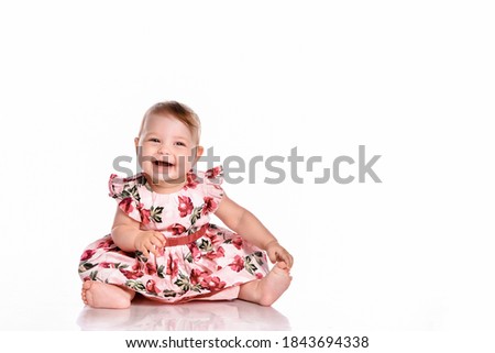 Portrait of a sweet infant girl isolated on white in studio. Girl dressed in a pink dress with flowers sits barefoot with a cute expressive face