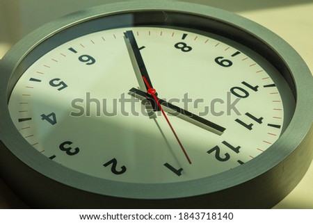 A Modern white wall clock in sunlight with shadows on the  clock face. Close up to a wall clock, with a red seconds pointer.