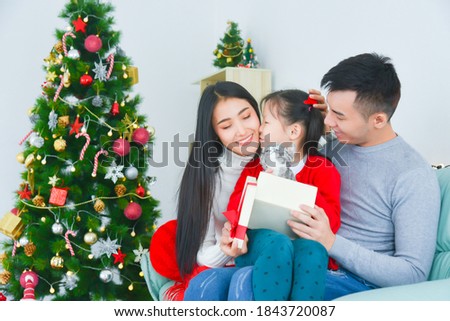 Asian mom and dad opening gift box with her daughter. Near the Christmas tree at home. Little girl kissing her mother,thanks for present.